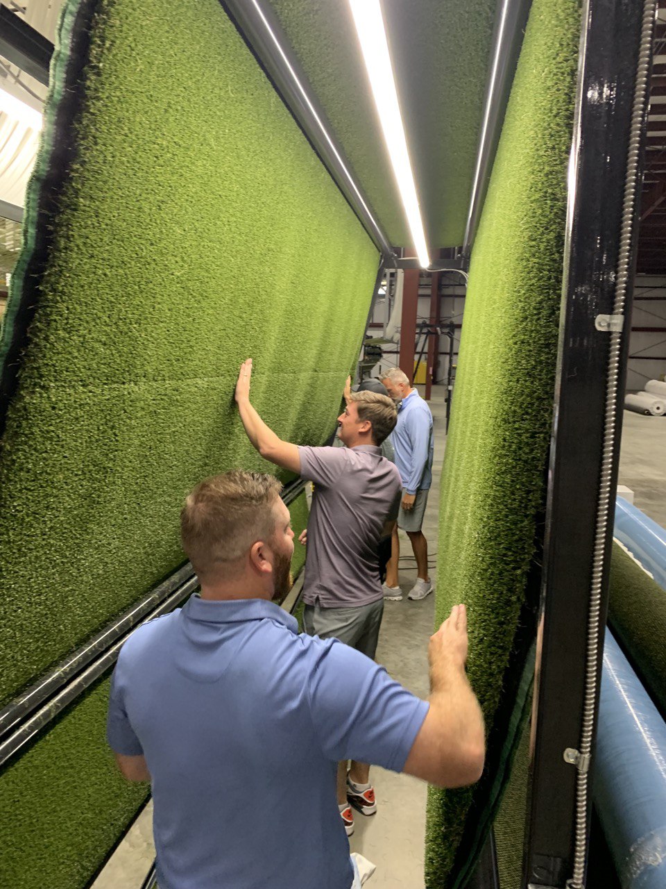 Workers inspecting large rolls of synthetic turf in an American artificial grass manufacturing facility. The quality control process ensures durability and precision in turf production for sports, landscaping, and commercial applications
