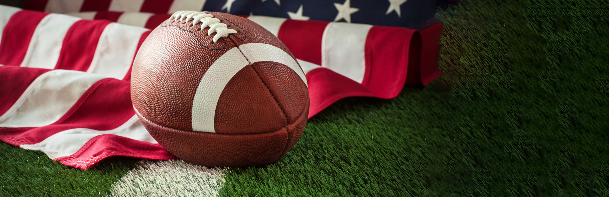 American football resting on a synthetic turf field with the U.S. flag in the background. The image represents patriotism, sportsmanship, and the use of high-quality artificial grass for professional and collegiate football games