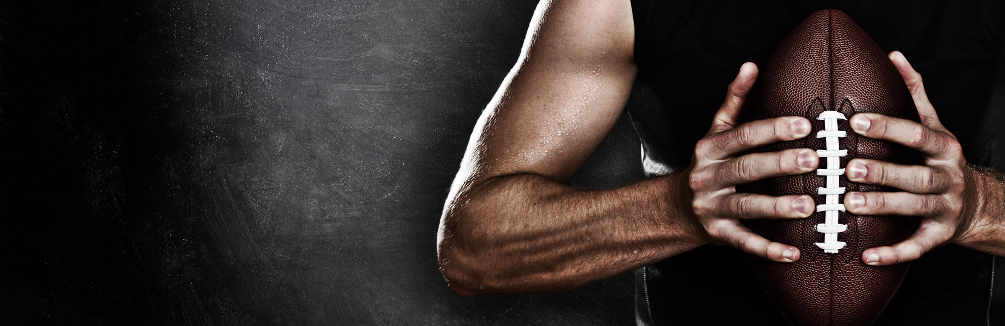 Close-up of a muscular athlete holding a football with both hands, emphasizing strength and grip, against a dark, dramatic background.