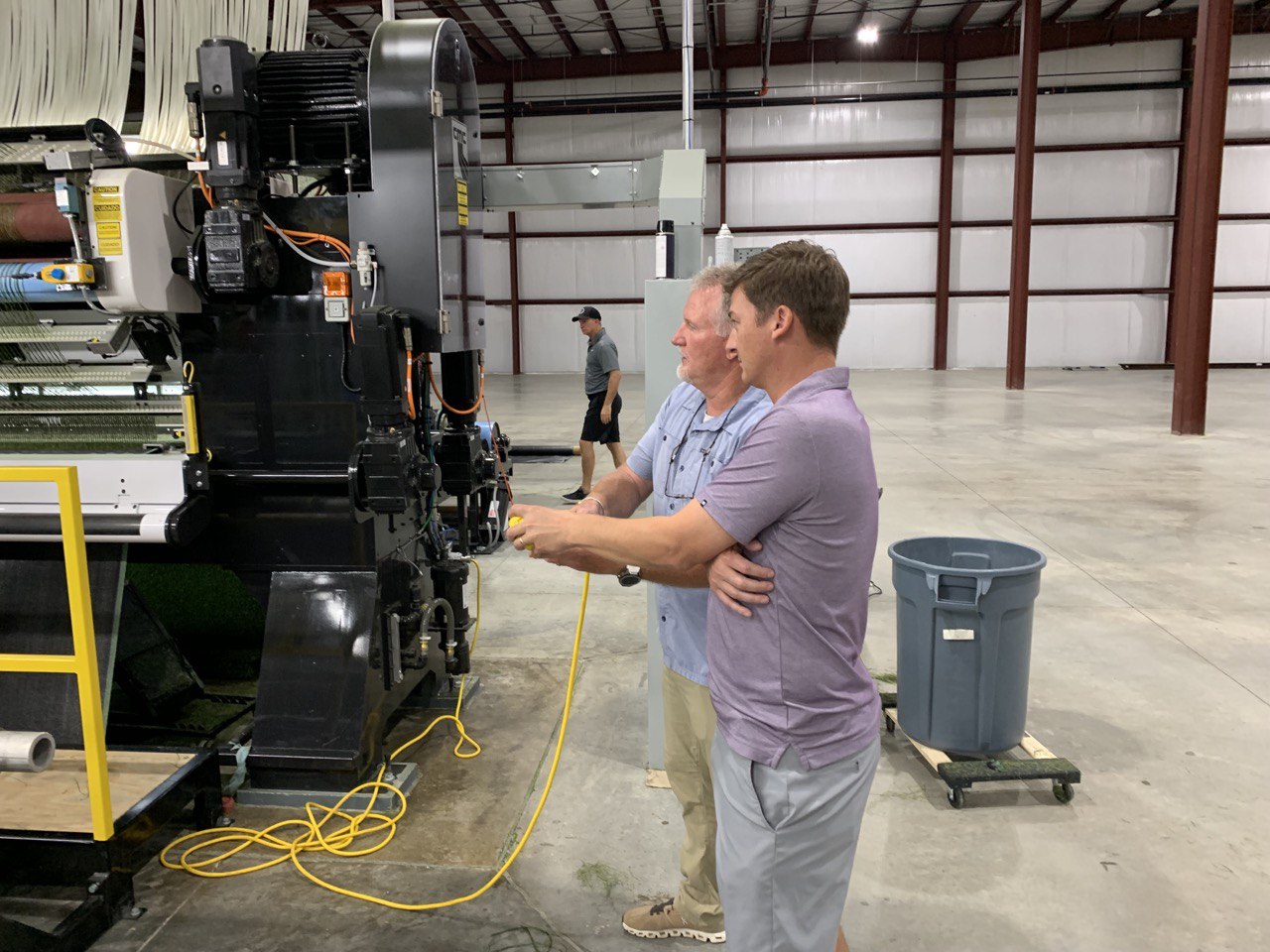 Engineers operating tufting machinery in an American synthetic turf manufacturing facility. The hands-on process highlights precision engineering and innovation in artificial grass production for sports, landscaping, and commercial applications