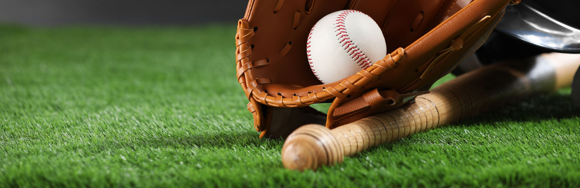A baseball glove with a ball resting on synthetic turf, next to a wooden bat, ready for gameplay.