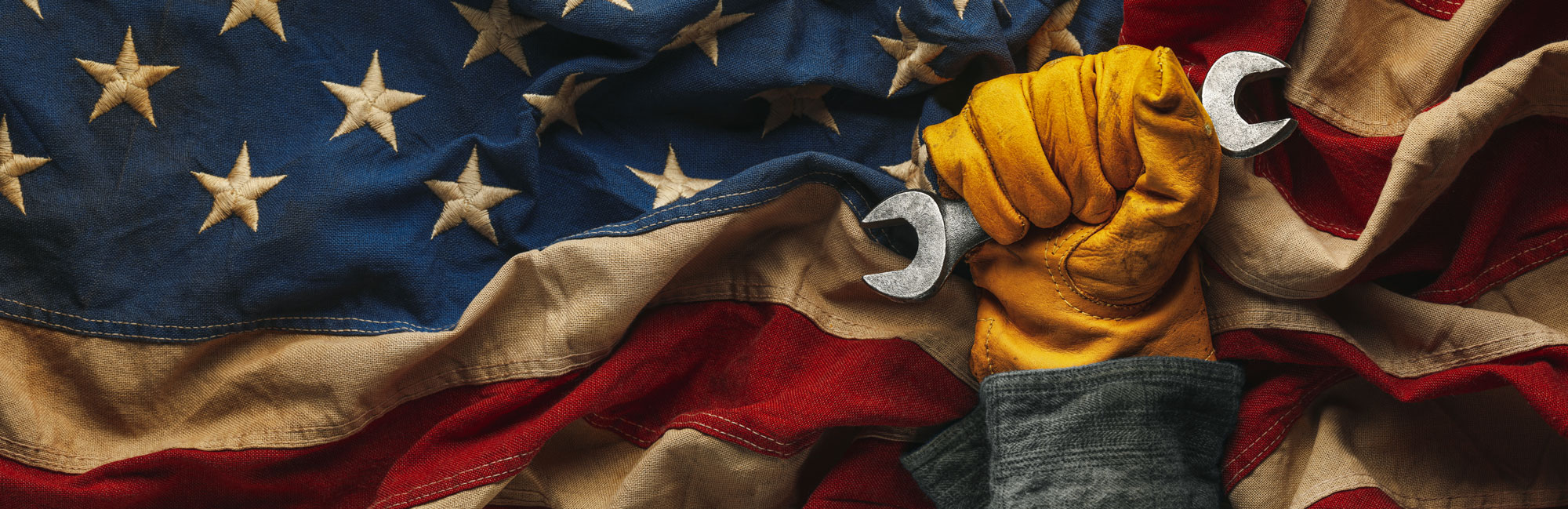 A worker gloved hand gripping wrenches, raised in front of an American flag. The image symbolizes strength, craftsmanship, and dedication to American manufacturing, including industries like synthetic turf production.