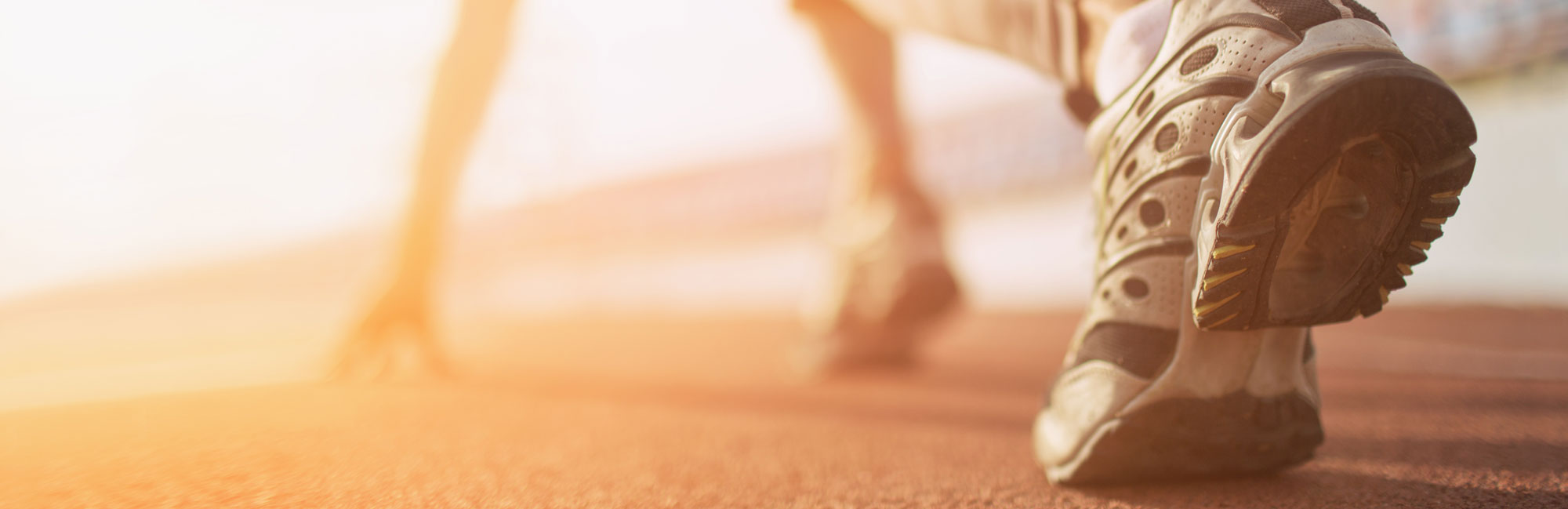 Close-up of a runners shoe on a professional track, preparing for a sprint. The background features warm sunlight, symbolizing endurance, speed, and high-performance sports training on synthetic turf and track surfaces.