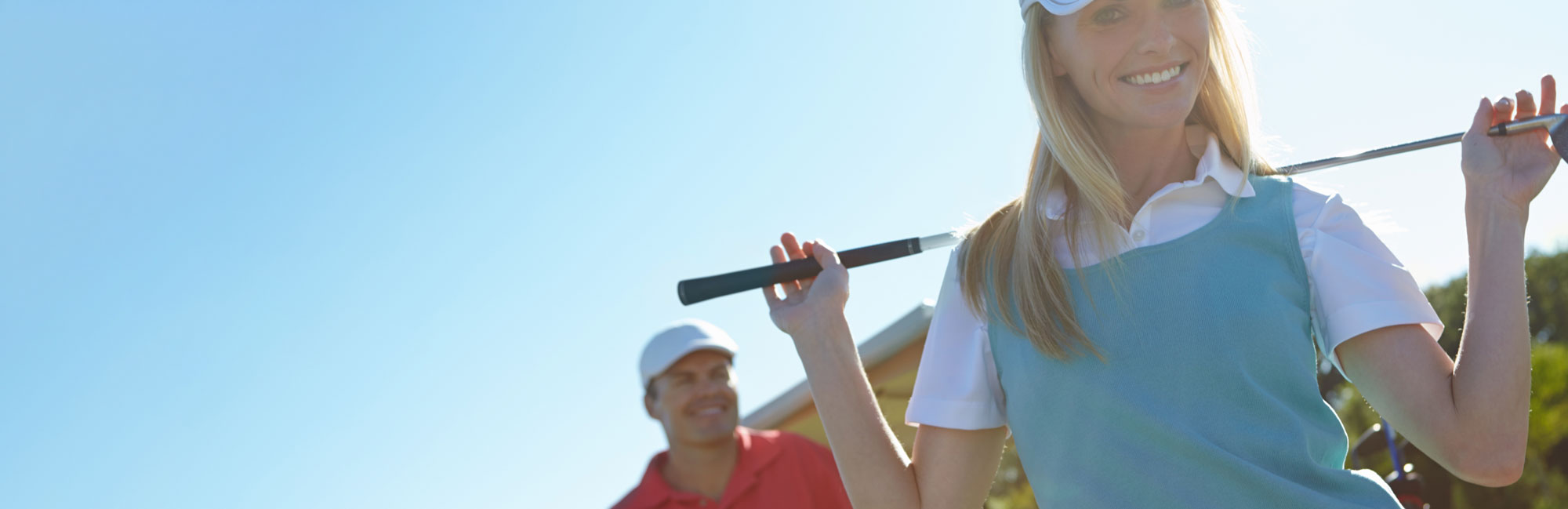 Female golfer holding a club over her shoulder, smiling with another golfer in the background under a clear sky.