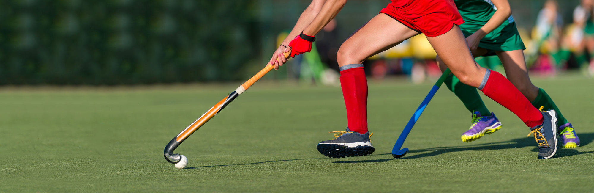 Field hockey players in red and green uniforms competing for the ball on synthetic turf, using their sticks for control.