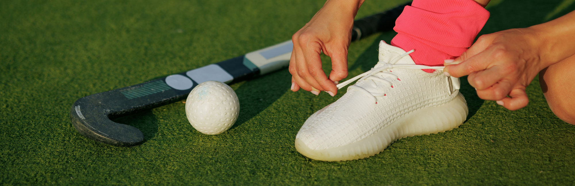 Close-up of a field hockey player tying shoelaces on synthetic turf, with a hockey stick and ball nearby.