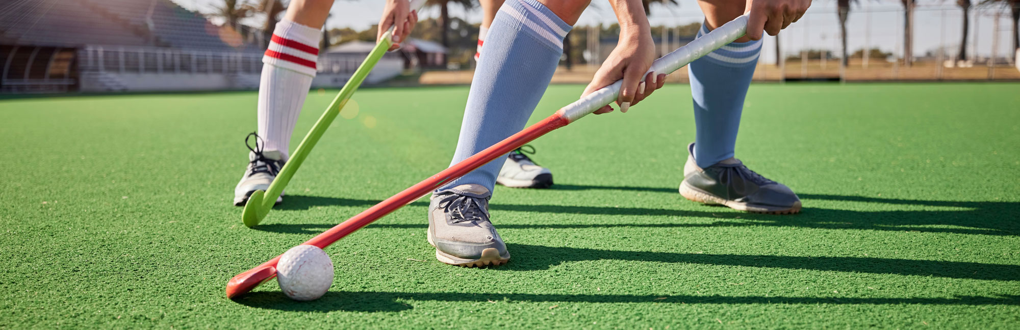 Field hockey players in action on synthetic turf, focusing on their sticks and ball movement under bright sunlight.
