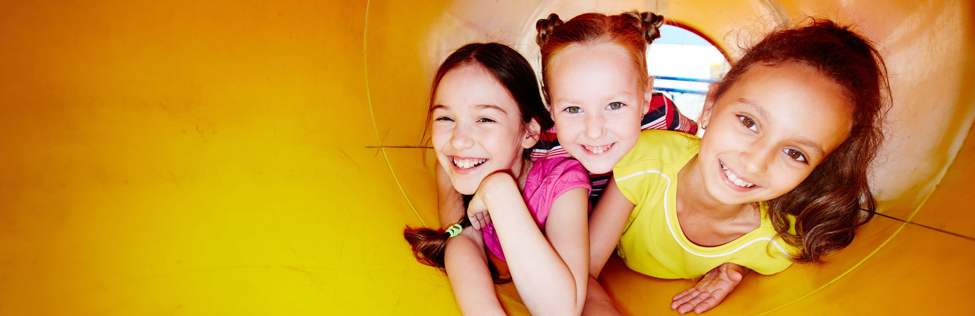 Three smiling young girls playing inside a yellow tunnel slide at a playground, looking joyful and excited.