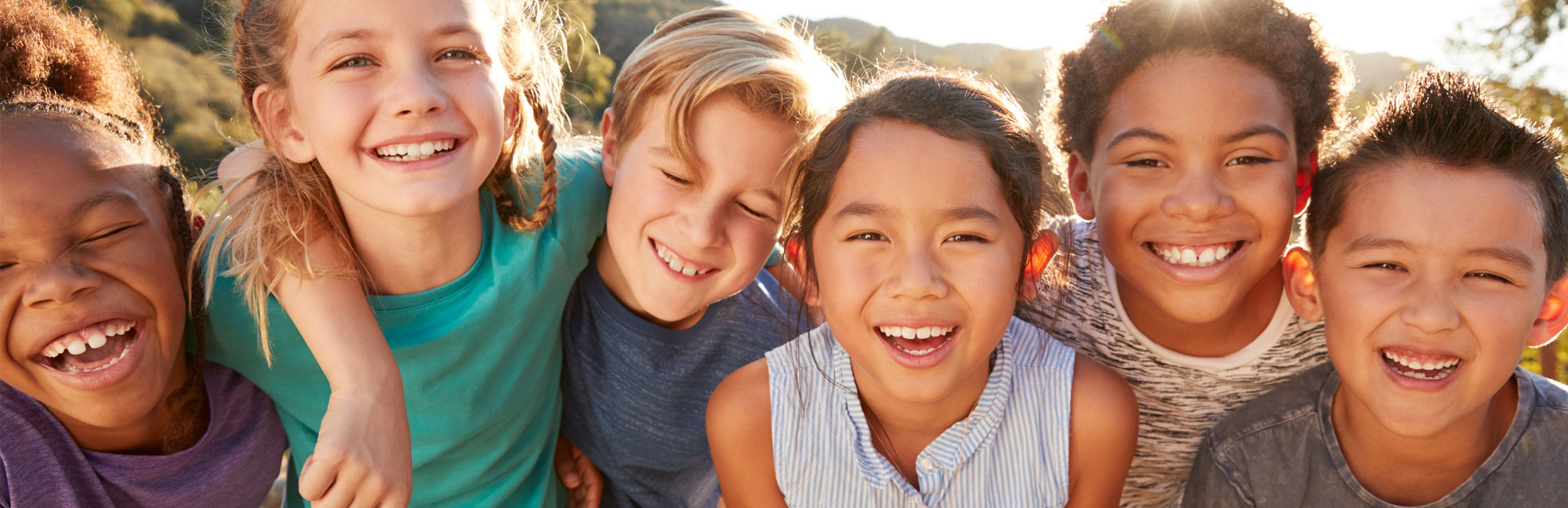 A group of diverse, happy children smiling and posing together outdoors in the sunlight.