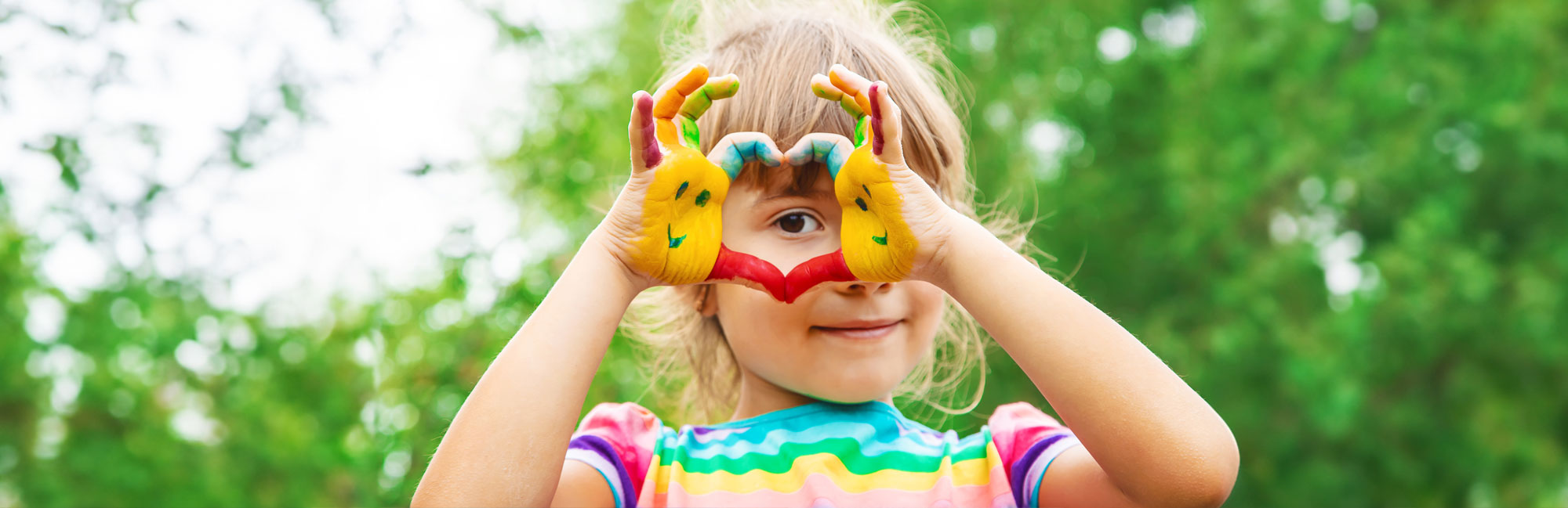 A cheerful young girl wearing a colorful striped shirt, playfully holding fruit-shaped glasses to her eyes while standing outdoors.