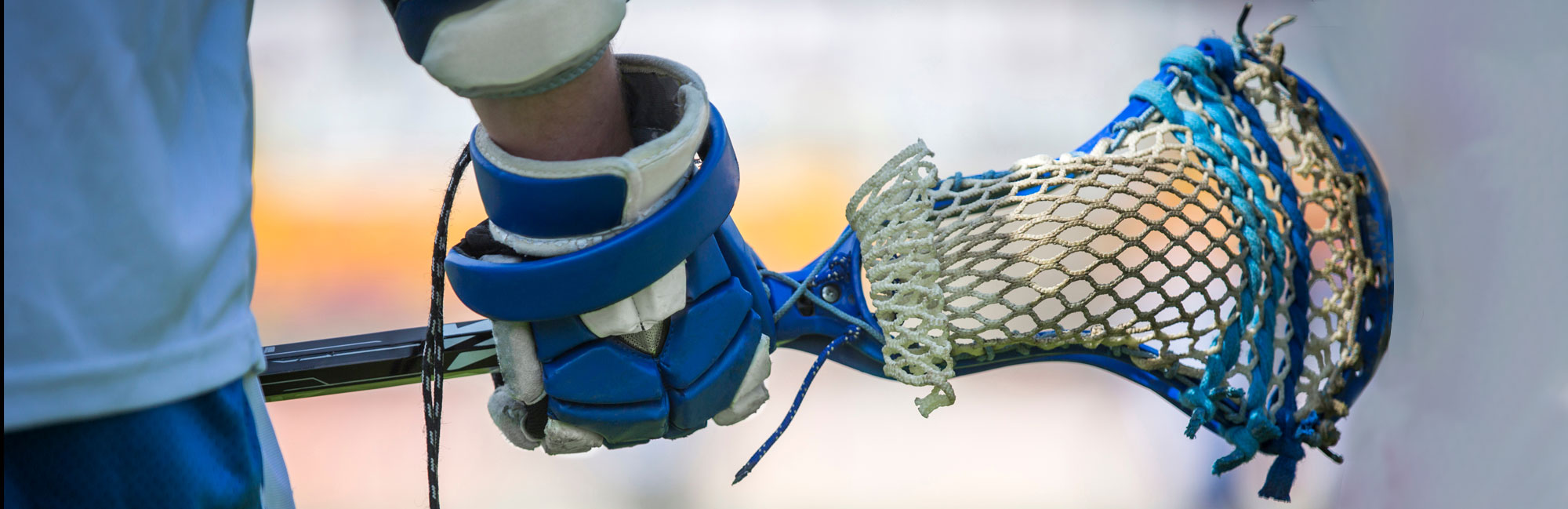 Close-up of a blue lacrosse stick held by a player in white and blue gear, with the netting ready to catch the ball.