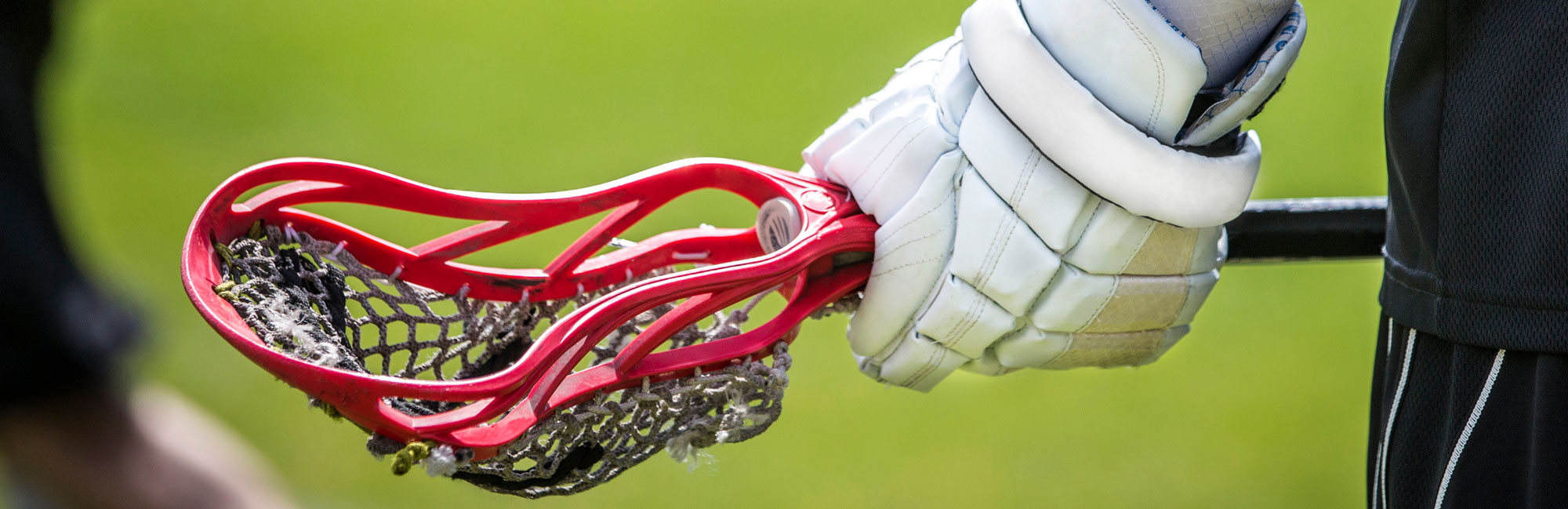 Close-up of a lacrosse stick with a red head, held by a player wearing white gloves, preparing for play on the field.