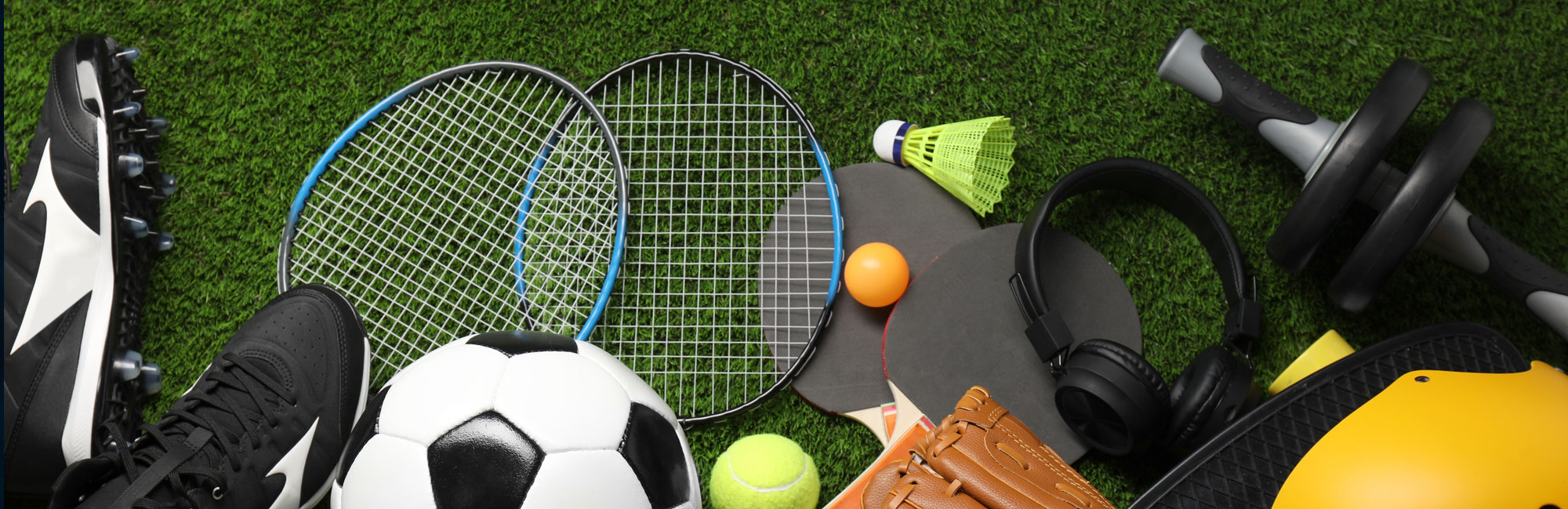 Various sports equipment, including soccer ball, tennis rackets, cleats, dumbbells, and gloves, laid out on synthetic turf.