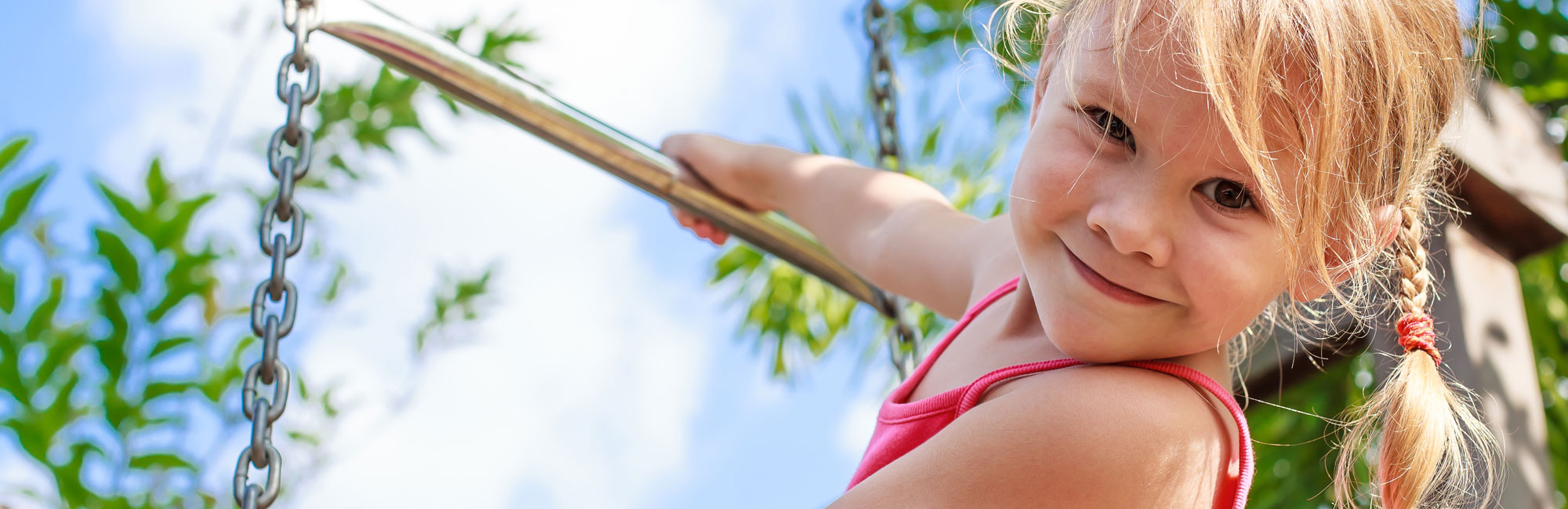 Smiling young girl swinging on a playground, holding onto a chain, with a bright blue sky and green foliage in the background.