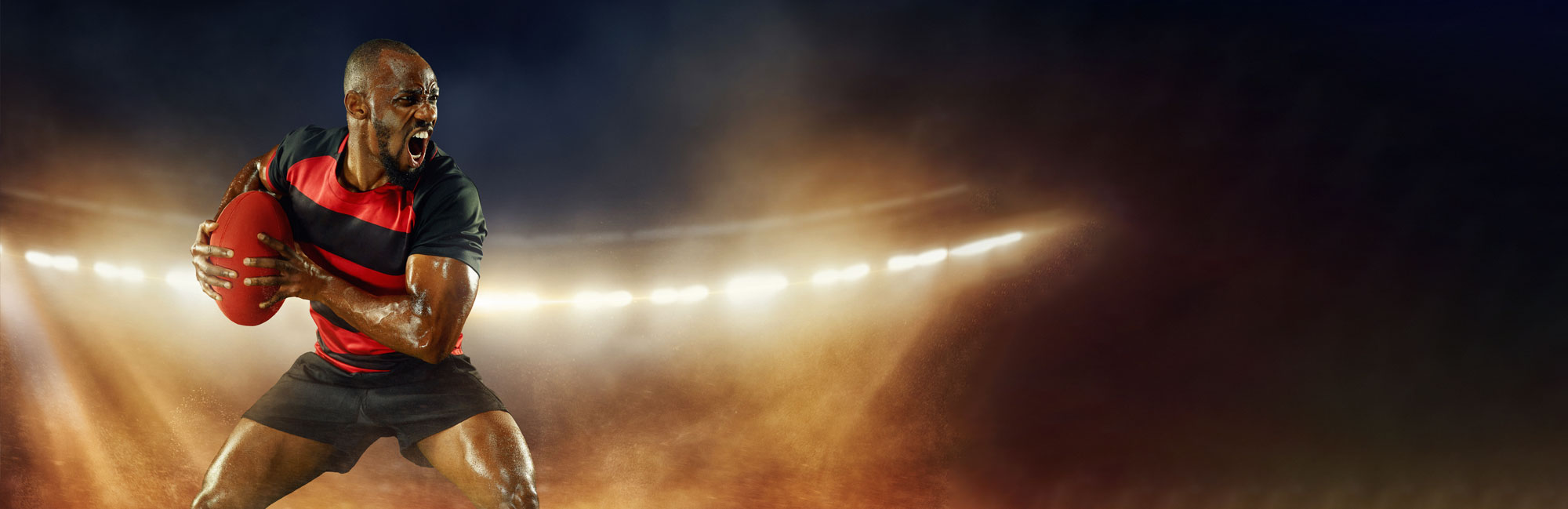 Rugby player gripping the ball in an intense action pose on a stadium field with synthetic turf. The dramatic lighting and dynamic movement highlight the power, speed, and durability of artificial grass for high-impact sports