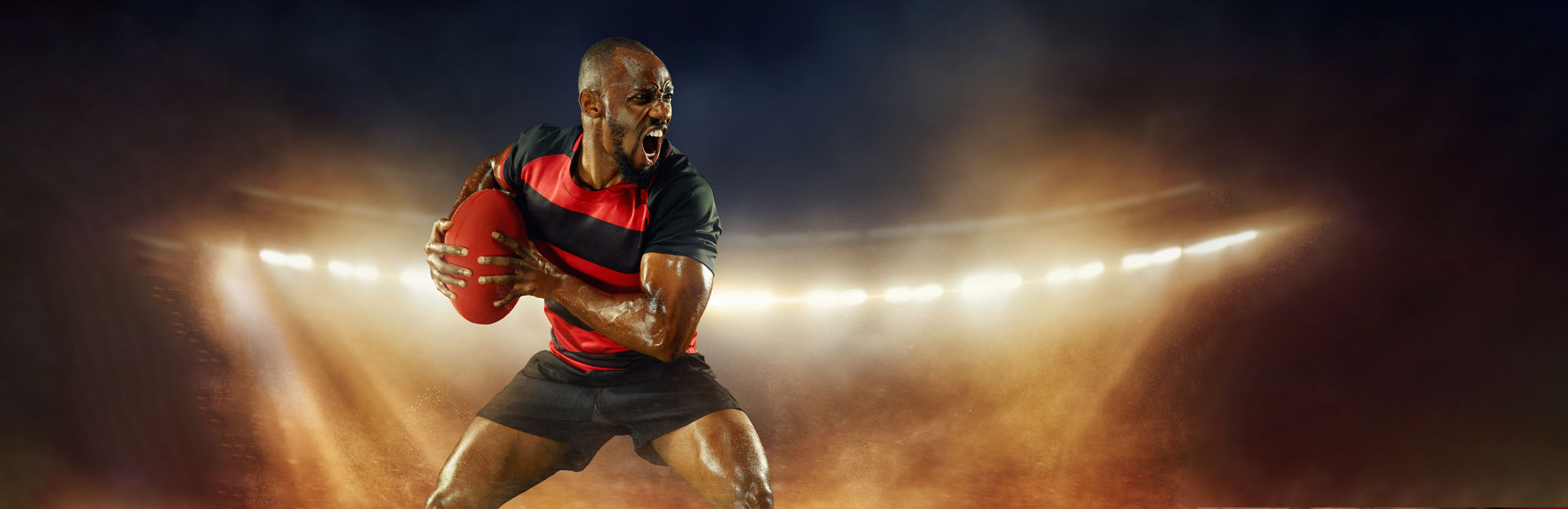 Rugby player gripping the ball in an intense action pose on a stadium field with synthetic turf. The dramatic lighting and dynamic movement highlight the power, speed, and durability of artificial grass for high-impact sports