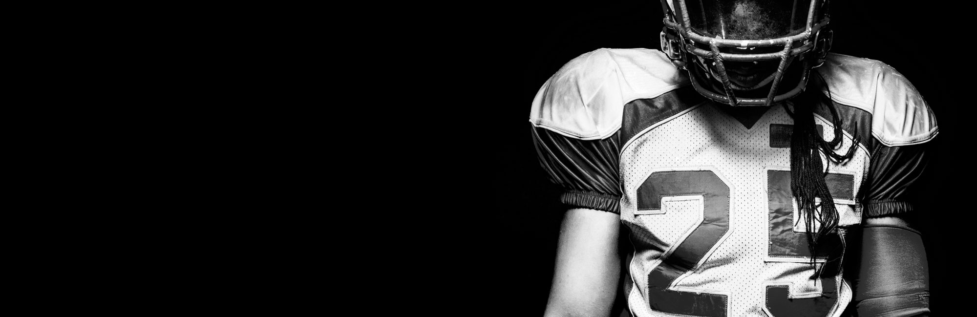 Black and white close-up of an American football player in full gear holding a ball. The dramatic contrast emphasizes strength, determination, and the role of high-quality synthetic turf in professional football games