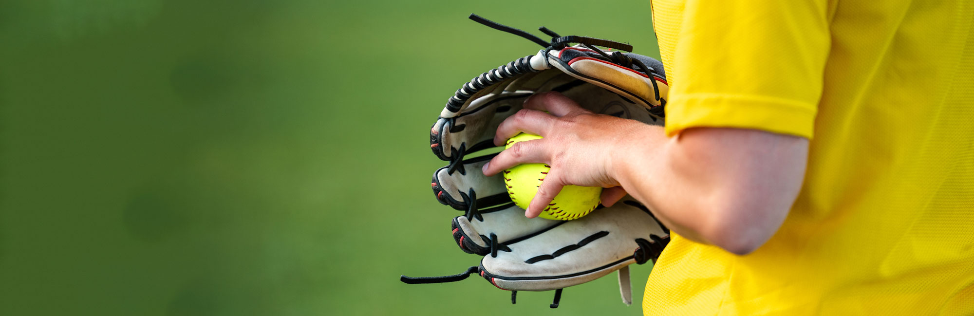 Softball player in a yellow jersey holding a bright yellow softball and a glove, preparing for the game on a green field.