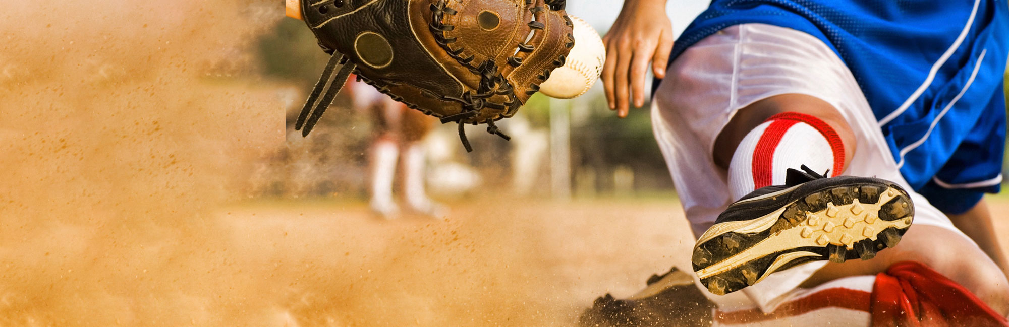 A softball player slides into base as a fielder reaches out with a glove to catch the ball, kicking up dust in the process.