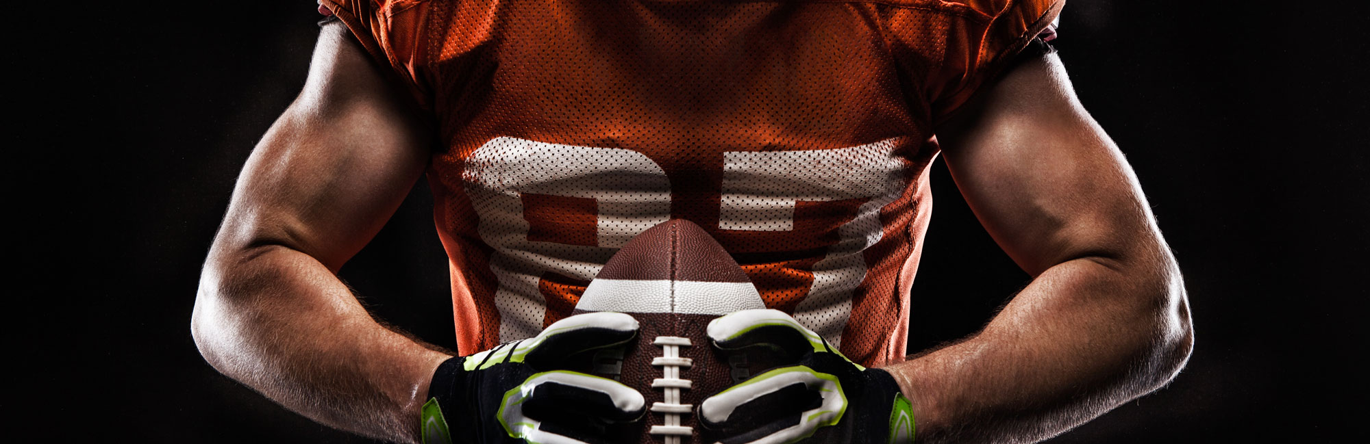 American football player in an orange jersey gripping a football, highlighting strength, determination, and the intensity of the game. The image represents professional sports played on high-quality synthetic turf fields