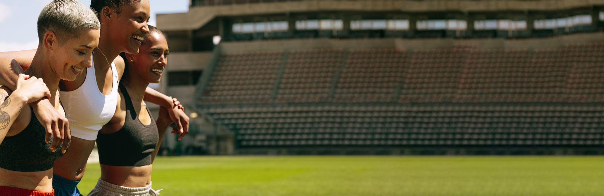 A group of female athletes embracing and celebrating on a professional synthetic turf field in a stadium. The scene highlights teamwork, resilience, and the high-quality artificial grass surface designed for peak sports performance.